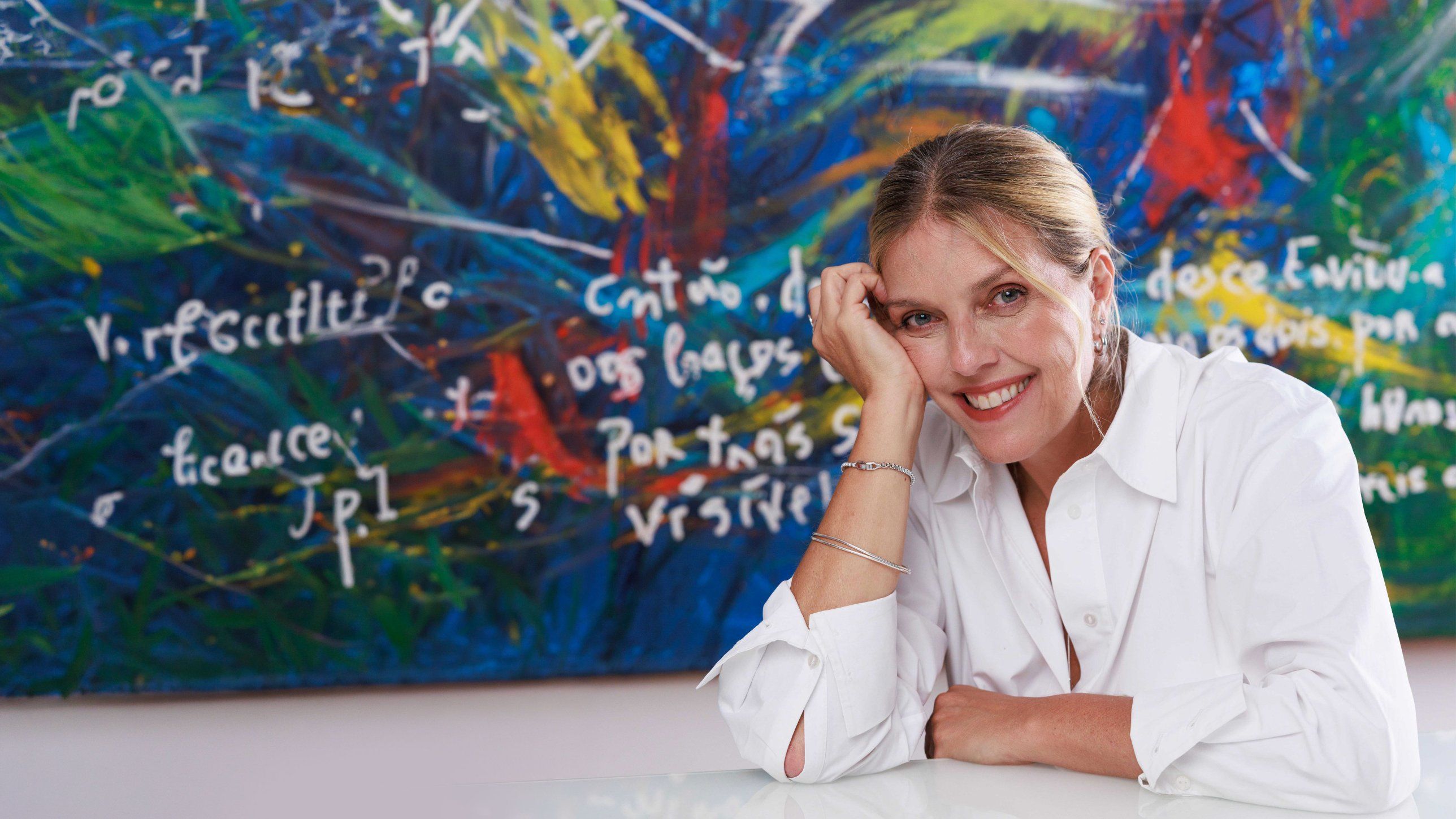 Patricia Tempski sorrindo diante de uma pintura em tons de azul e verde
